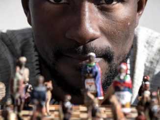 Teddy Kossoko, assis devant un jeu d’échecs avec des pions à l’effigie de personnages africains.