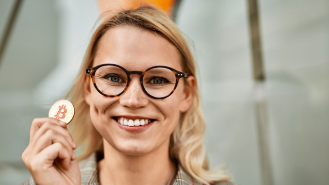 Une jeune femme blonde, sourire aux lèvres, brandissant un jeton physique de Bitcoin.