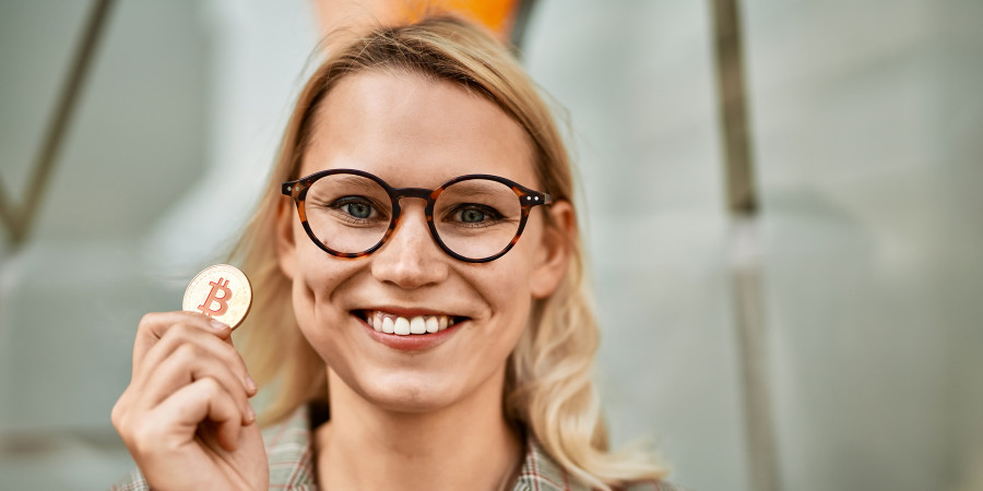 Une jeune femme blonde, sourire aux lèvres, brandissant un jeton physique de Bitcoin.